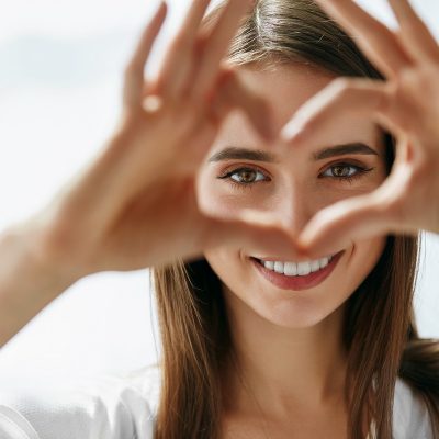 Beautiful Happy Woman Showing Love Sign Near Eyes. Healthy Eyes And Vision. Portrait Of Beautiful Happy Woman Holding Heart Shaped Hands Near Eyes. Closeup Of Smiling Girl With Healthy Skin Showing Love Sign. Eyecare. High Resolution Image